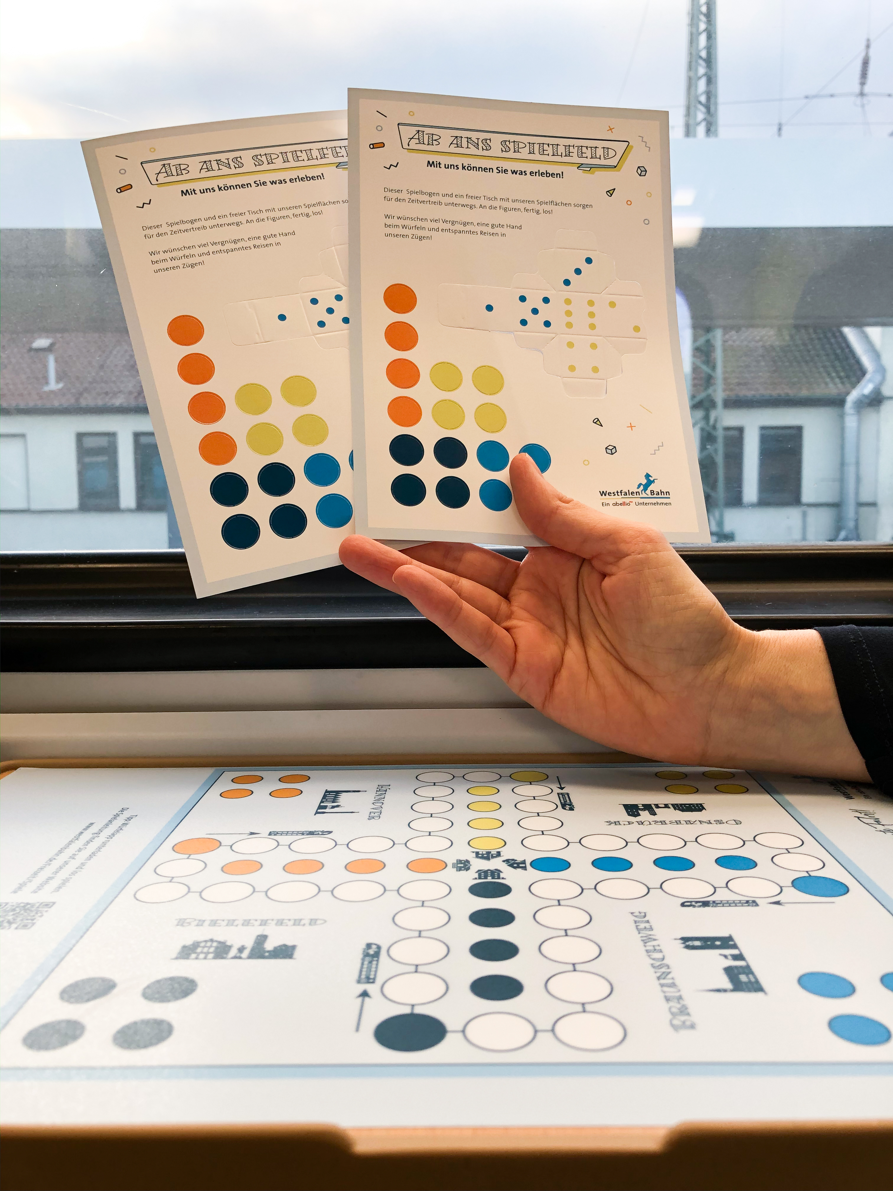 Eine Hand hält die Spielanleitungen zum Brettspiel "Mensch-Ärgere-Dich-Nicht" in die Kamera, darunter ist das Spiel auf einen Zugtisch geklebt.
