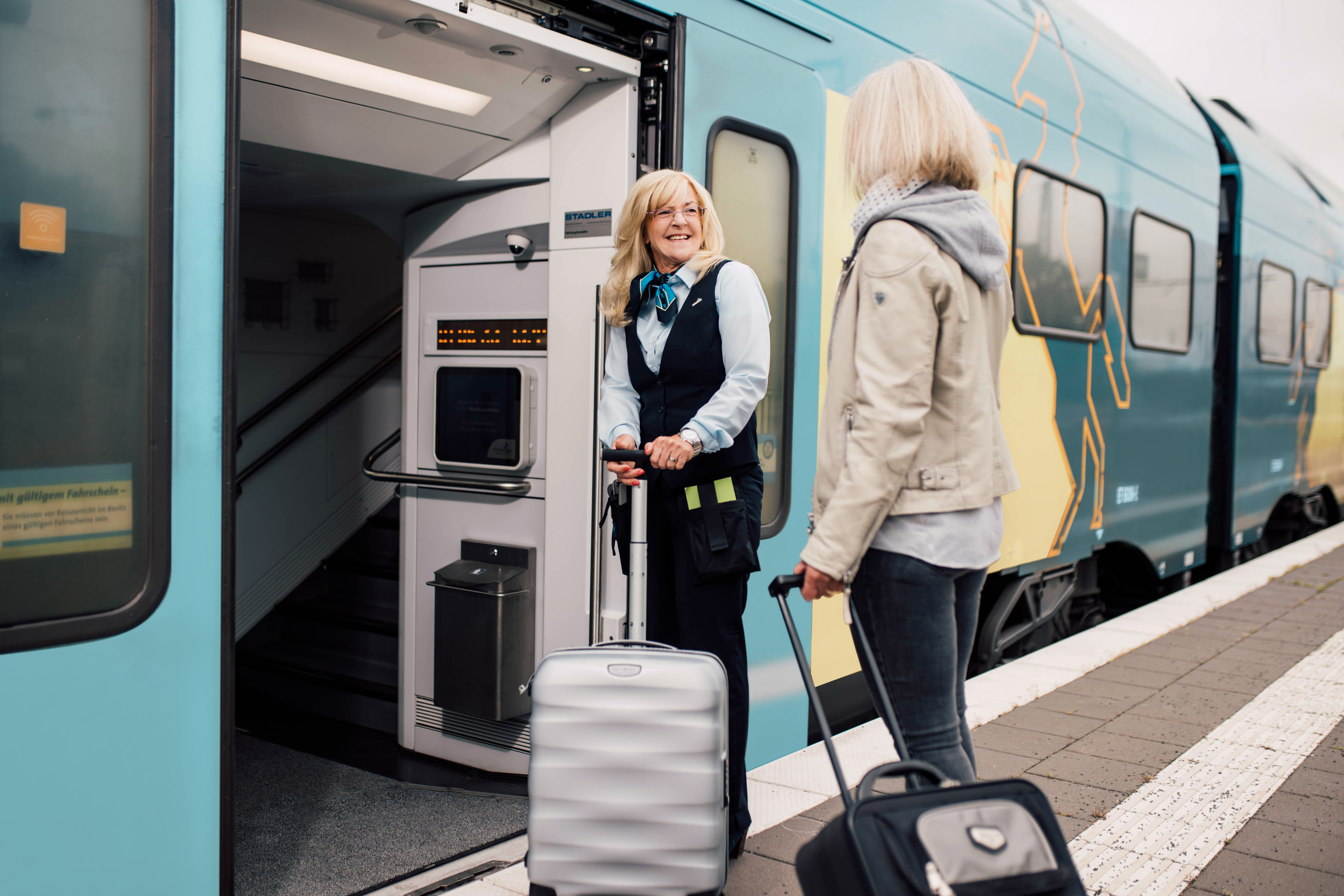 Eine Kundenbetreuerin hilft einer Fahrgästin mit Koffer, in einen Zug der WestfalenBahn einzusteigen.